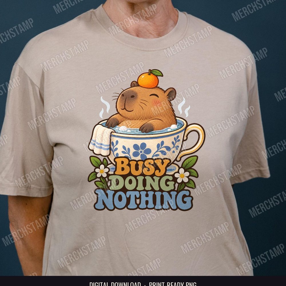 Busy Doing Nothing PNG design shown on a tan t-shirt, a relaxed capybara soaking in a floral teacup with an orange on its head above the words Busy Doing Nothing