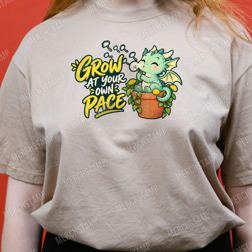 Grow At Your Own Pace PNG design shown on a tan t-shirt, a green baby dragon in a terracotta pot blowing dandelion seeds beside the words Grow At Your Own Pace
